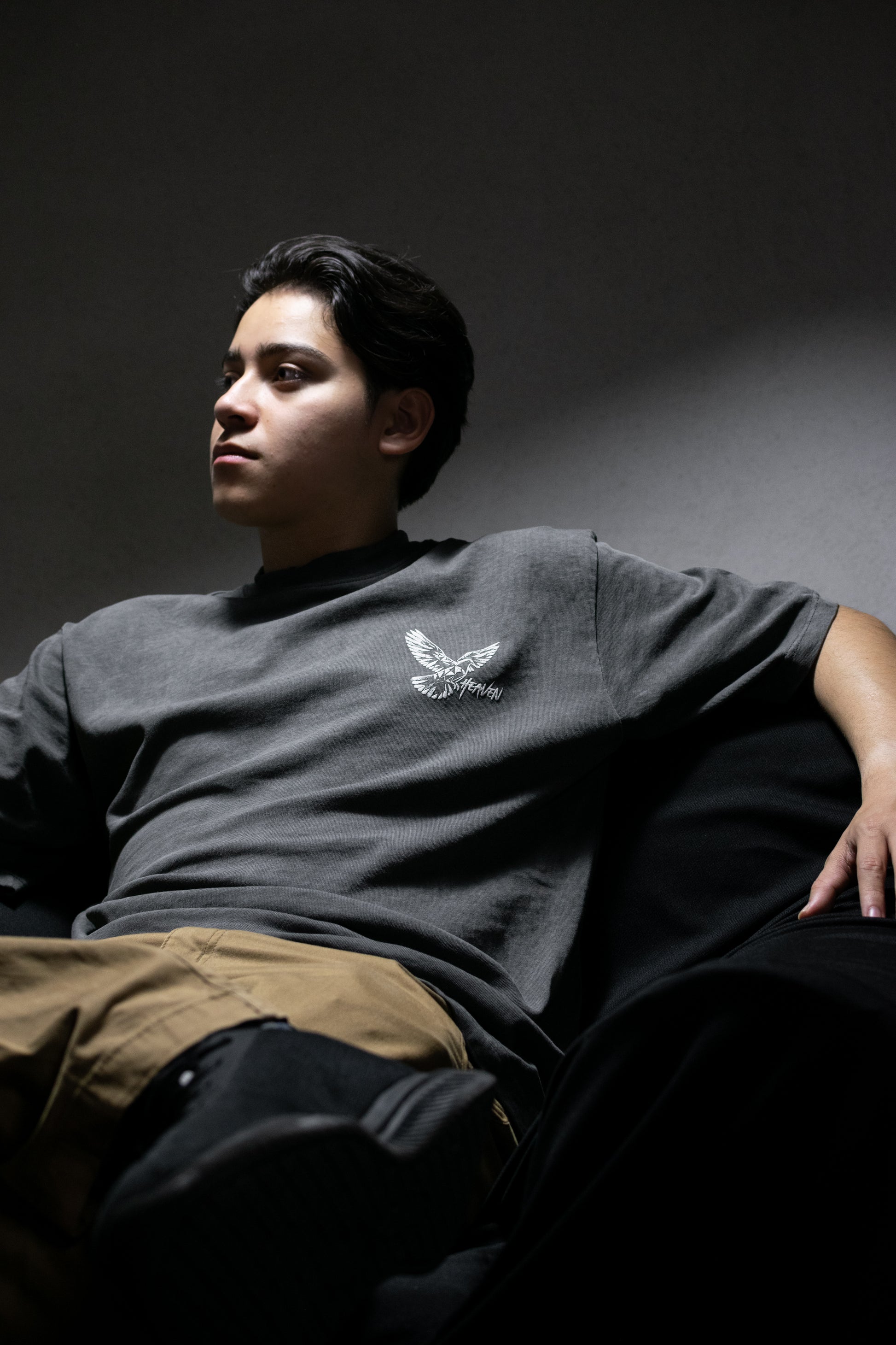Person wearing a gray t-shirt with a logo, sitting against a dark background.